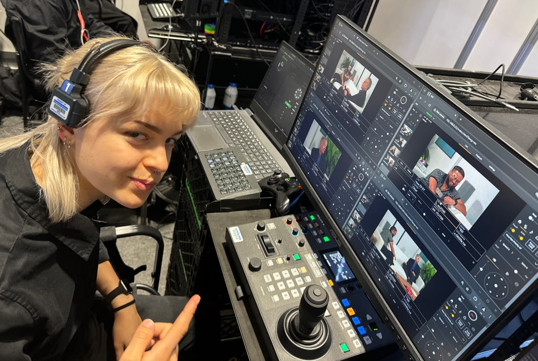 Emmy sits at a camera operating station (table with knobs and buttons, computer monitors with camera displays).